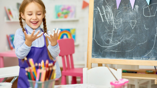 Portrait of cute little girl enjoying art and craft lesson in development school and smiling happily looking at hands covered in paint