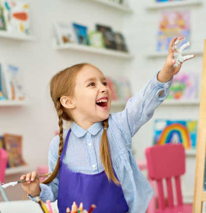 Portrait of adorable little girl enjoying art and craft lesson in development school and smiling happily with hands in paint